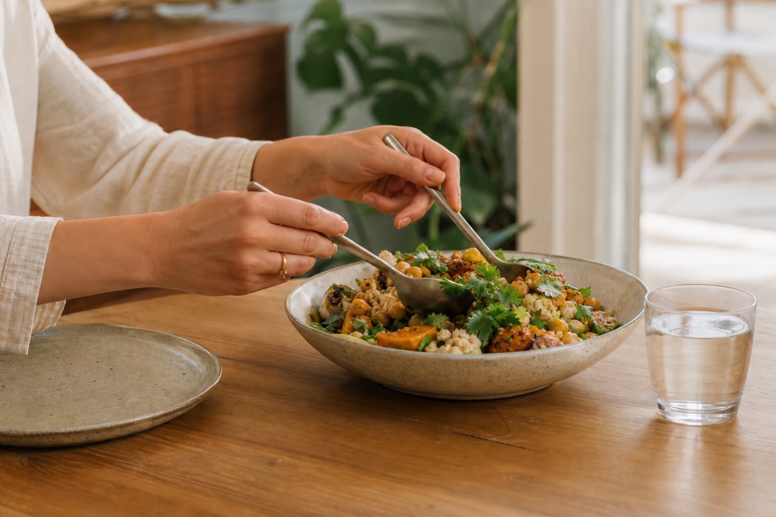 Hände nehmen sich entspannt von einer bunten Gemüse-Bowl an einem hellen Holztisch, daneben ein Glas Wasser.